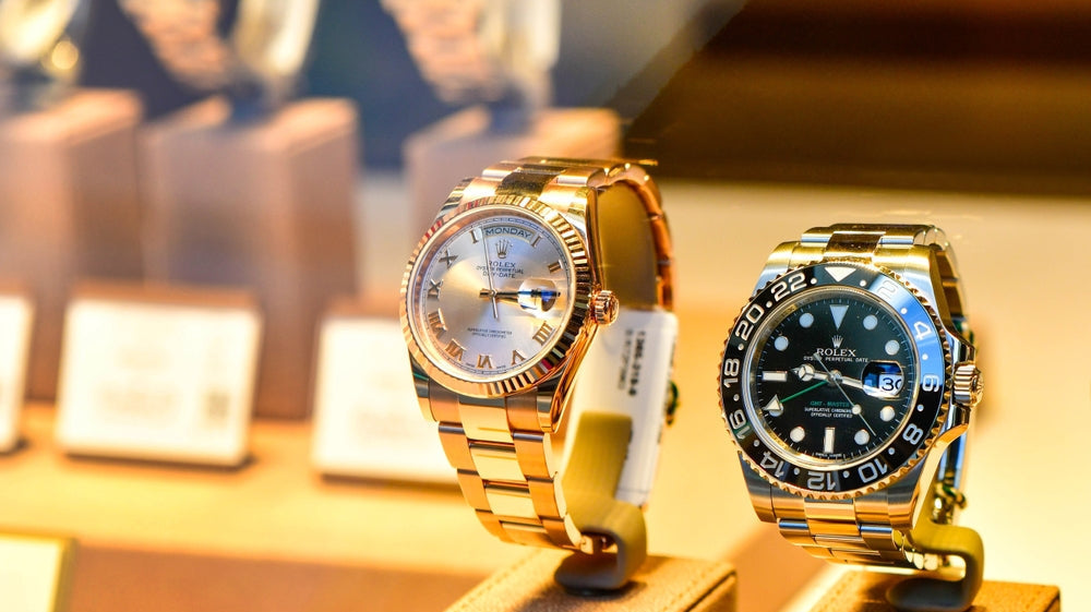 Two luxury Rolex watches on display: a rose gold Rolex Day-Date with Roman numeral dial (left) and a two-tone Rolex GMT-Master II with black dial and Cerachrom bezel (right), showcased in a high-end jewelry store