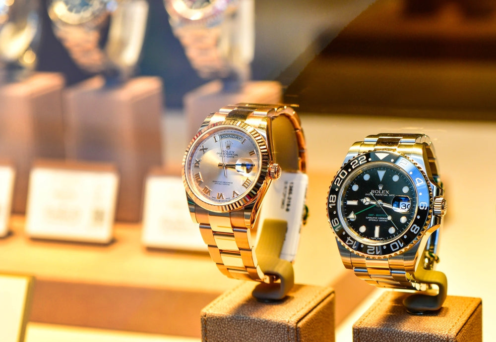 Two luxury Rolex watches on display: a rose gold Rolex Day-Date with Roman numeral dial (left) and a two-tone Rolex GMT-Master II with black dial and Cerachrom bezel (right), showcased in a high-end jewelry store